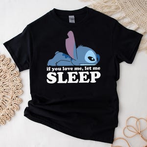 K&ouml;nnte beinhalten: Schwarzes T-Shirt mit einer blauen Stitch-Figur aus Lilo & Stitch. Die Figur liegt mit dem Text "If you love me, let me SLEEP" in wei&szlig; gedruckt.