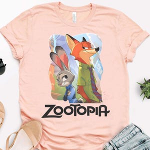 May include: A light pink t-shirt featuring the Zootopia logo in black, with a graphic of Nick Wilde and Judy Hopps. Nick is an orange fox wearing a green shirt and tie, and Judy is a gray rabbit in a blue uniform.
