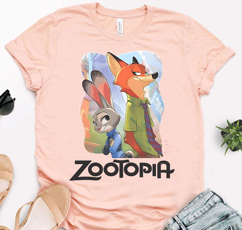 May include: A light pink t-shirt featuring the Zootopia logo in black, with a graphic of Nick Wilde and Judy Hopps. Nick is an orange fox wearing a green shirt and tie, and Judy is a gray rabbit in a blue uniform.