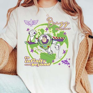 Puede incluir: Camiseta color crema con un gr&aacute;fico de Buzz Lightyear en un traje espacial, volando frente a un globo verde. El texto "Buzz Lightyear" y "to infinity and beyond" tambi&eacute;n se muestran en morado y amarillo.