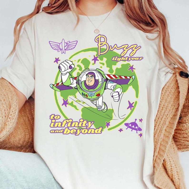Puede incluir: Camiseta color crema con un gr&aacute;fico de Buzz Lightyear en un traje espacial, volando frente a un globo verde. El texto "Buzz Lightyear" y "to infinity and beyond" tambi&eacute;n se muestran en morado y amarillo.