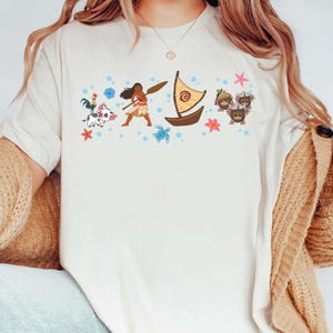 May include: Cream-coloured t-shirt with a colourful Polynesian-themed graphic. The design includes a boat, a woman with a paddle, and various cartoon characters. The shirt has a crew neck and short sleeves.
