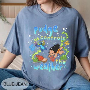 Puede incluir: Camiseta azul vaquero con un gr&aacute;fico de Stitch y Lilo bajo el agua, con el texto "Pudge controls the weather". El dise&ntilde;o incluye personajes de dibujos animados, burbujas y coral.