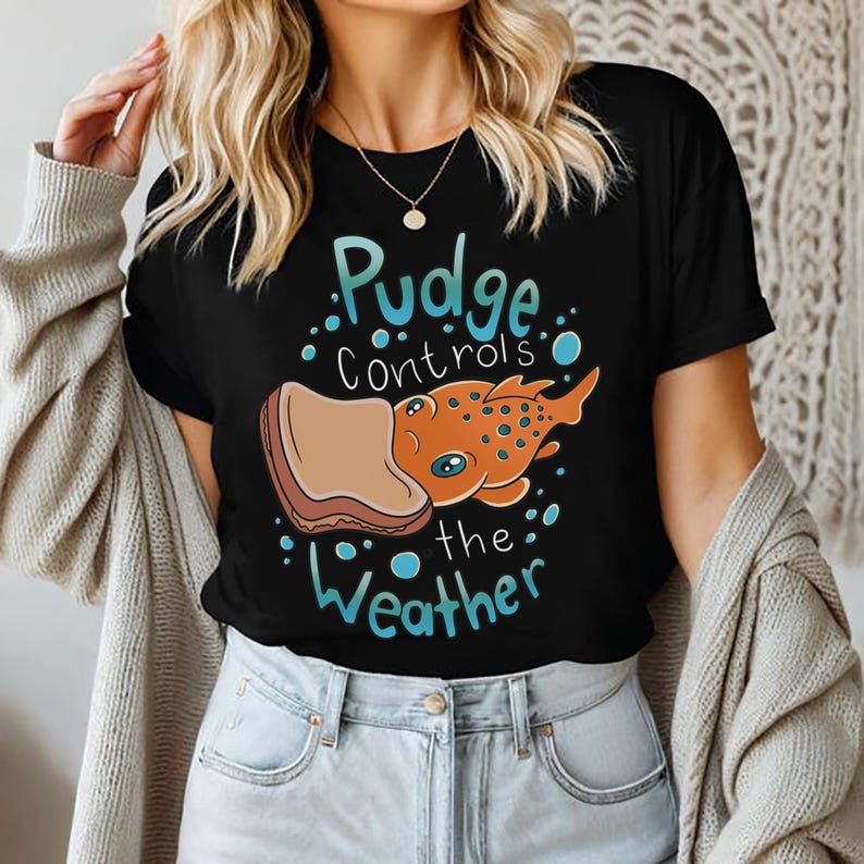 May include: Black t-shirt with a cartoon illustration of a fish eating a slice of bread. The text "Pudge controls the weather" is printed on the shirt.