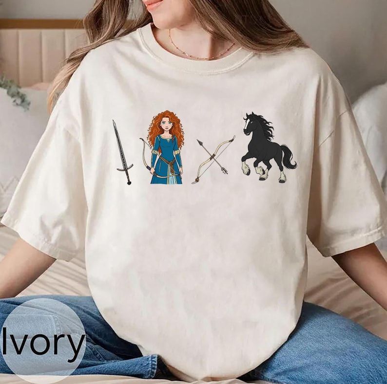 May include: Ivory-colored t-shirt featuring a cartoon illustration of a sword, a bow and arrow, a woman in a blue dress, and a black horse. The word "Ivory" is printed at the bottom of the shirt.