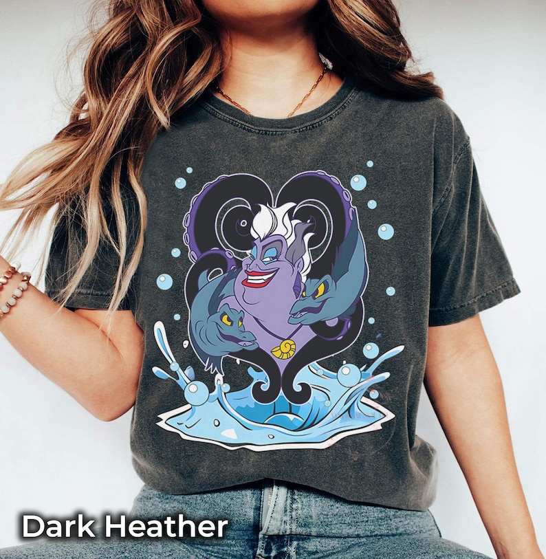 May include: Dark heather gray t-shirt featuring a graphic of Ursula from The Little Mermaid, with Flotsam and Jetsam, and water splash details. The text "Dark Heather" is at the bottom of the image.