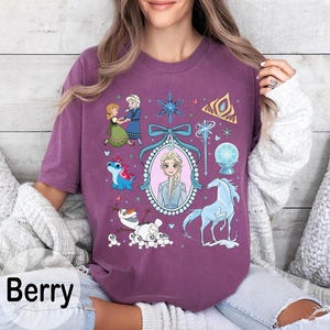 May include: Berry-coloured t-shirt featuring a collage of characters and elements from a popular animated film. The design includes illustrations of two princesses, a snowman, a blue horse, and other related graphics. The word "Berry" is printed at the bottom.