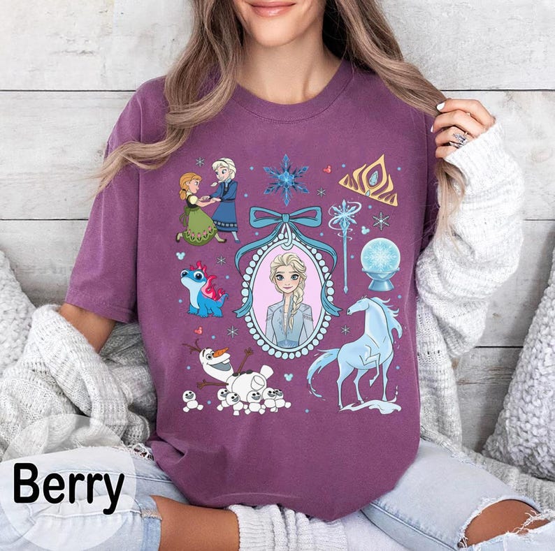 May include: Berry-coloured t-shirt featuring a collage of characters and elements from a popular animated film. The design includes illustrations of two princesses, a snowman, a blue horse, and other related graphics. The word "Berry" is printed at the bottom.