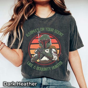 May include: Dark heather gray t-shirt featuring a graphic of a bounty hunter with the text "Karma's on your scent like a bounty hunter." The design includes a circular sunset background. The shirt is a comfortable, casual wear.