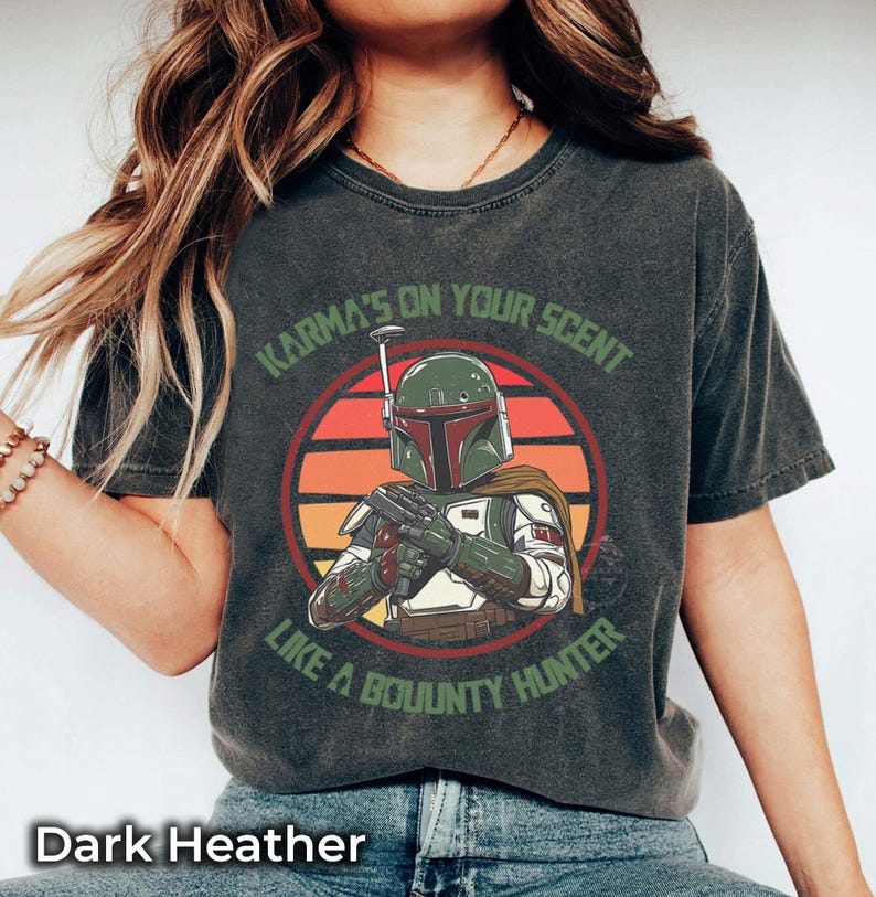 May include: Dark heather gray t-shirt featuring a graphic of a bounty hunter with the text "Karma's on your scent like a bounty hunter." The design includes a circular sunset background. The shirt is a comfortable, casual wear.