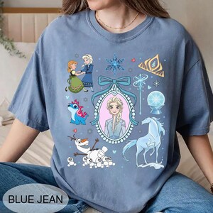 May include: A blue jean coloured t-shirt with cartoon images of characters from the film Frozen. The shirt features Elsa, Anna, Olaf, and other elements from the film. The text "BLUE JEAN" is printed at the bottom.