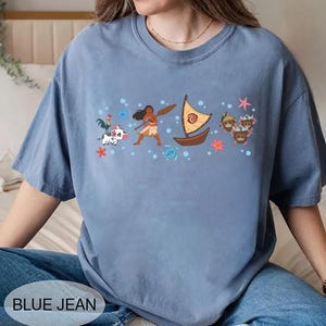 May include: A blue jean coloured t-shirt with a graphic of a boat, characters, and the text "BLUE JEAN". The graphic includes a boat with a yellow sail, a woman with a paddle, and other cartoon characters. The shirt is a solid colour.