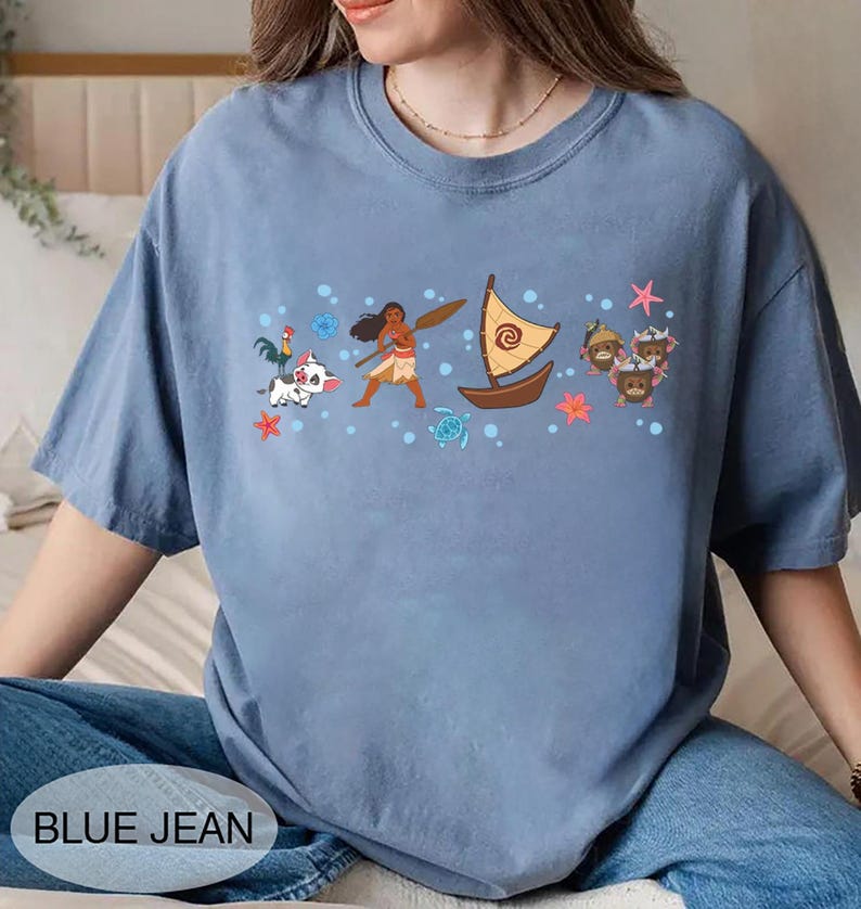 May include: A blue jean coloured t-shirt with a graphic of a boat, characters, and the text "BLUE JEAN". The graphic includes a boat with a yellow sail, a woman with a paddle, and other cartoon characters. The shirt is a solid colour.