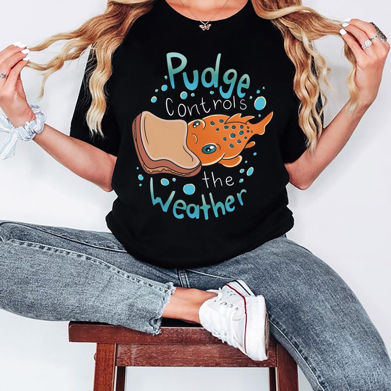 May include: A black t-shirt with a graphic of a cartoon fish with a slice of bread on its head. The text on the shirt reads "Pudge controls the weather".