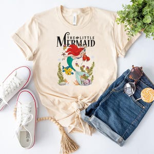 Op de afbeelding: Een licht beige T-shirt met een afbeelding van Ariel, de Kleine Zeemeermin, die zwemt met Flounder. De tekst "The Little Mermaid" is in zwart boven de afbeelding gedrukt.