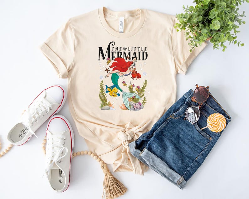 Op de afbeelding: Een licht beige T-shirt met een afbeelding van Ariel, de Kleine Zeemeermin, die zwemt met Flounder. De tekst "The Little Mermaid" is in zwart boven de afbeelding gedrukt.