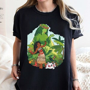 Puede incluir: Camiseta negra con un gr&aacute;fico colorido de una mujer polinesia, una diosa verde, un gallo y un cerdo. El dise&ntilde;o est&aacute; ambientado en un tel&oacute;n de fondo tropical con palmeras y flores. La obra de arte es vibrante y detallada, perfecta para los fan&aacute;ticos de la pel&iacute;cula animada.