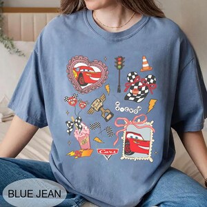 May include: Blue jean colored t-shirt featuring a Cars movie graphic. The design includes Lightning McQueen, racing flags, and other themed illustrations. The shirt has a crew neck and short sleeves.