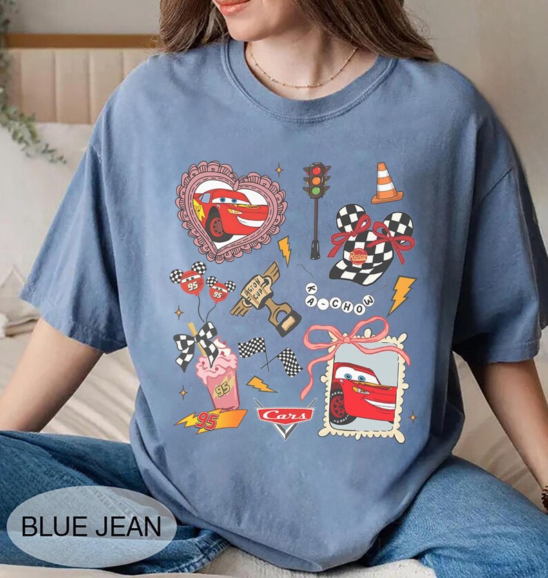 May include: Blue jean colored t-shirt featuring a Cars movie graphic. The design includes Lightning McQueen, racing flags, and other themed illustrations. The shirt has a crew neck and short sleeves.
