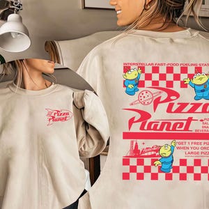 May include: Beige sweatshirt with a red Pizza Planet logo on the front and back. The back features the text "INTERSTELLAR FAST-FOOD FUELING STATION" and cartoon aliens. The design includes the words "PIZZAS, SALADS, SNACKS, BEVERAGES" and "GET 1 FREE PIZZA WHEN YOU ORDER LARGE PIZZAS."