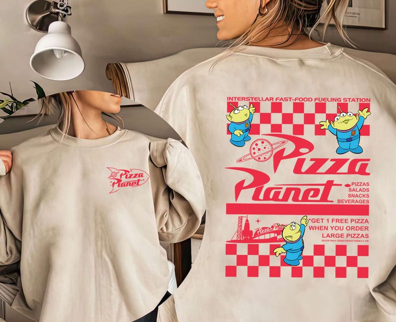 May include: Beige sweatshirt with a red Pizza Planet logo on the front and back. The back features the text "INTERSTELLAR FAST-FOOD FUELING STATION" and cartoon aliens. The design includes the words "PIZZAS, SALADS, SNACKS, BEVERAGES" and "GET 1 FREE PIZZA WHEN YOU ORDER LARGE PIZZAS."