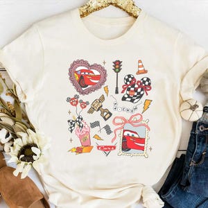 May include: Cream-colored t-shirt featuring a graphic design with elements from the movie Cars. The design includes a red race car, checkered flags, and other racing-themed illustrations. The word "Cars" is also visible.