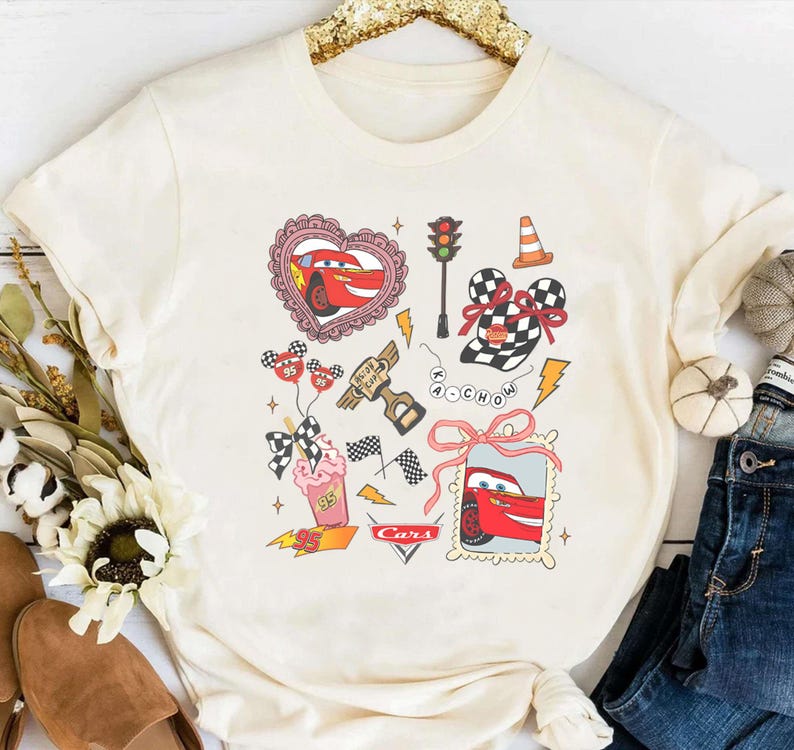 May include: Cream-colored t-shirt featuring a graphic design with elements from the movie Cars. The design includes a red race car, checkered flags, and other racing-themed illustrations. The word "Cars" is also visible.