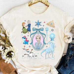 May include: Cream-coloured t-shirt with a vibrant graphic of characters and elements from a popular animated film. The design includes illustrations of a princess, a horse, snowflakes, and other related imagery. The shirt is made of a soft material.