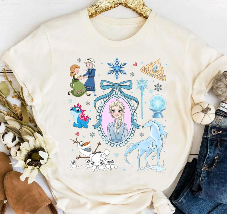 May include: Cream-coloured t-shirt with a vibrant graphic of characters and elements from a popular animated film. The design includes illustrations of a princess, a horse, snowflakes, and other related imagery. The shirt is made of a soft material.