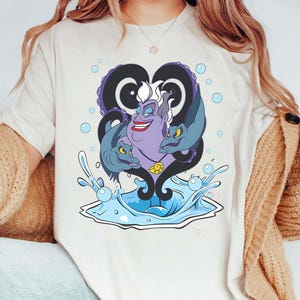 May include: A cream-colored t-shirt featuring a cartoon illustration of Ursula from The Little Mermaid. The design includes Ursula's face, tentacles, and two eels, with water and bubbles. The artwork is in shades of purple, blue, and white.