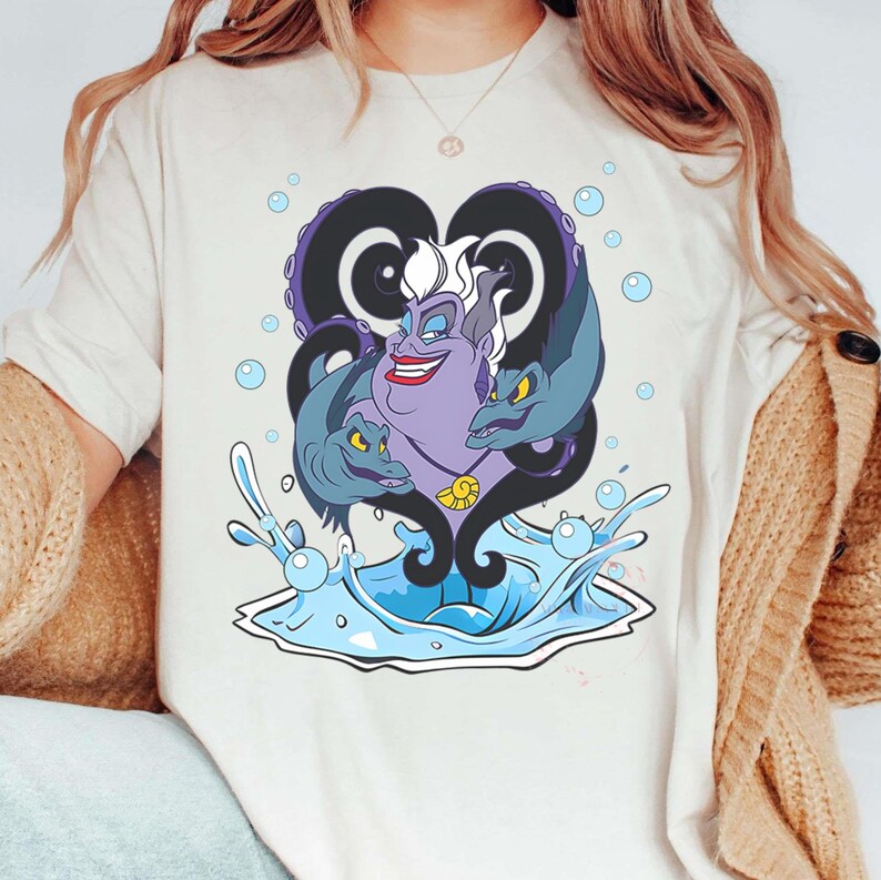 May include: A cream-colored t-shirt featuring a cartoon illustration of Ursula from The Little Mermaid. The design includes Ursula's face, tentacles, and two eels, with water and bubbles. The artwork is in shades of purple, blue, and white.