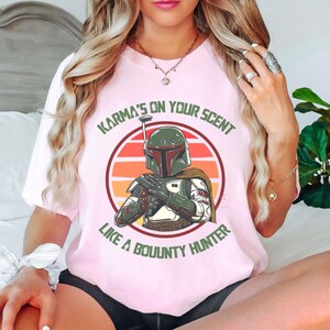 May include: A light pink t-shirt featuring a graphic of a bounty hunter in a circle with a sunset background. The text "Karma's on your scent like a bounty hunter" is printed above and below the graphic. The shirt is a crew neck.