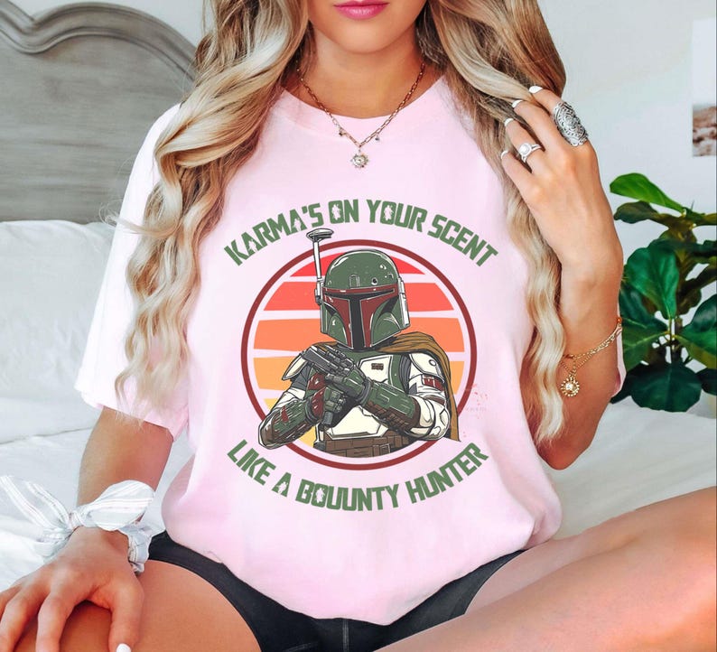 May include: A light pink t-shirt featuring a graphic of a bounty hunter in a circle with a sunset background. The text "Karma's on your scent like a bounty hunter" is printed above and below the graphic. The shirt is a crew neck.
