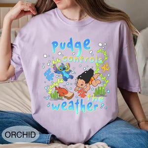 Puede incluir: Camiseta de color lila claro con un dise&ntilde;o de dibujos animados. El dise&ntilde;o presenta el texto "pudge controls the weather" con los personajes Stitch y Lilo, adem&aacute;s de criaturas marinas y burbujas. La camiseta est&aacute; etiquetada "ORCHID" en la parte inferior.