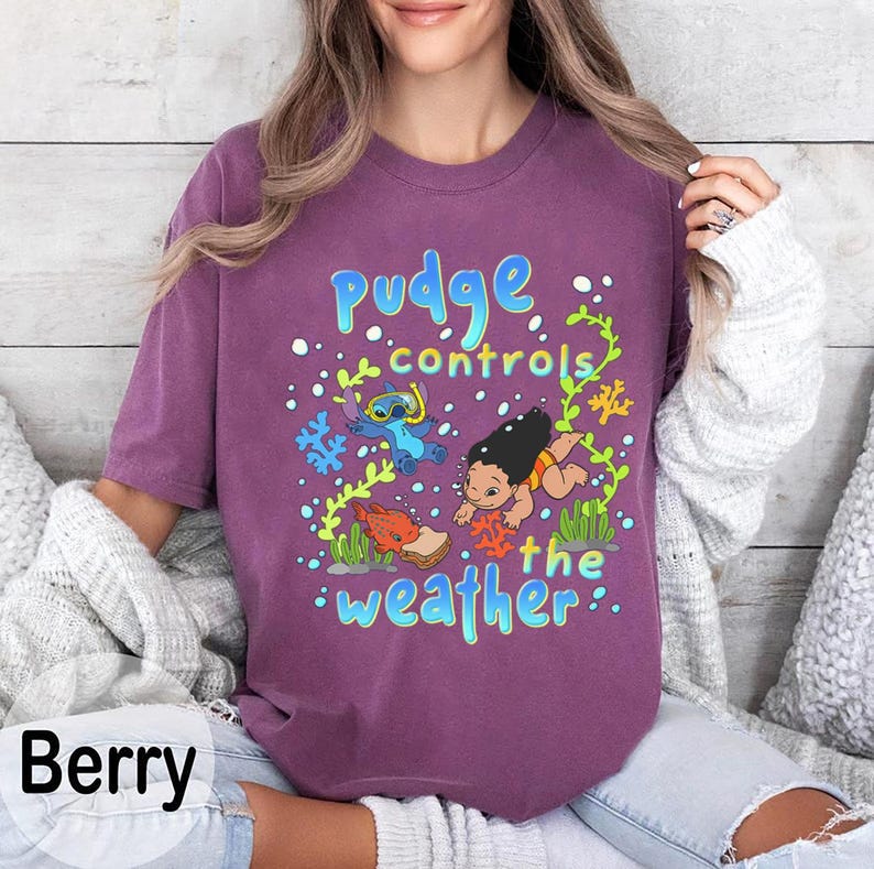 Puede incluir: Camiseta de color bayas con un gr&aacute;fico colorido que dice "Pudge controla el clima". El dise&ntilde;o incluye personajes de dibujos animados y elementos acu&aacute;ticos en azul, verde y naranja. Una prenda de vestir informal.