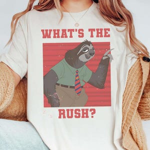 May include: Cream-colored t-shirt featuring a cartoon sloth wearing a green shirt and a red and blue striped tie. The text "WHAT'S THE RUSH?" is printed above and below the sloth illustration in a red, distressed font.