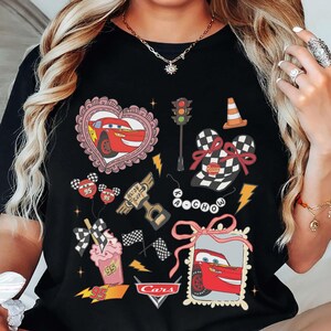 May include: Black t-shirt featuring various Cars movie-themed graphics. Designs include Lightning McQueen in a heart, a traffic light, a cone, checkered flag elements, and the Cars logo. The shirt has a crew neck.