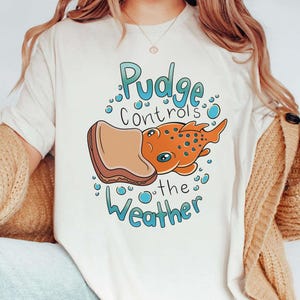 May include: White t-shirt with a cartoon illustration of a fish with a peanut butter and jelly sandwich on its head. The text "Pudge Controls the Weather" is printed on the shirt.