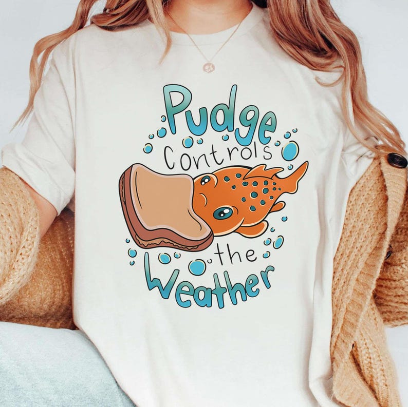 May include: White t-shirt with a cartoon illustration of a fish with a peanut butter and jelly sandwich on its head. The text "Pudge Controls the Weather" is printed on the shirt.