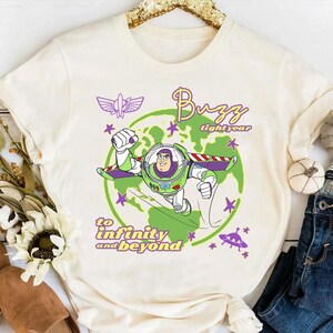 Puede incluir: Camiseta color crema con un gr&aacute;fico de Buzz Lightyear con un traje espacial verde y morado, con las palabras "Buzz Lightyear" y "hasta el infinito y m&aacute;s all&aacute;". El dise&ntilde;o incluye un planeta verde, estrellas y una nave espacial morada.