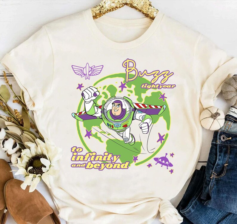 Puede incluir: Camiseta color crema con un gr&aacute;fico de Buzz Lightyear con un traje espacial verde y morado, con las palabras "Buzz Lightyear" y "hasta el infinito y m&aacute;s all&aacute;". El dise&ntilde;o incluye un planeta verde, estrellas y una nave espacial morada.