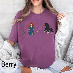 May include: Berry-colored t-shirt featuring a cartoon illustration of a character with red hair, a sword, bow and arrow, and a black horse. The shirt has a relaxed fit and short sleeves. The word "Berry" is printed in white at the bottom left.