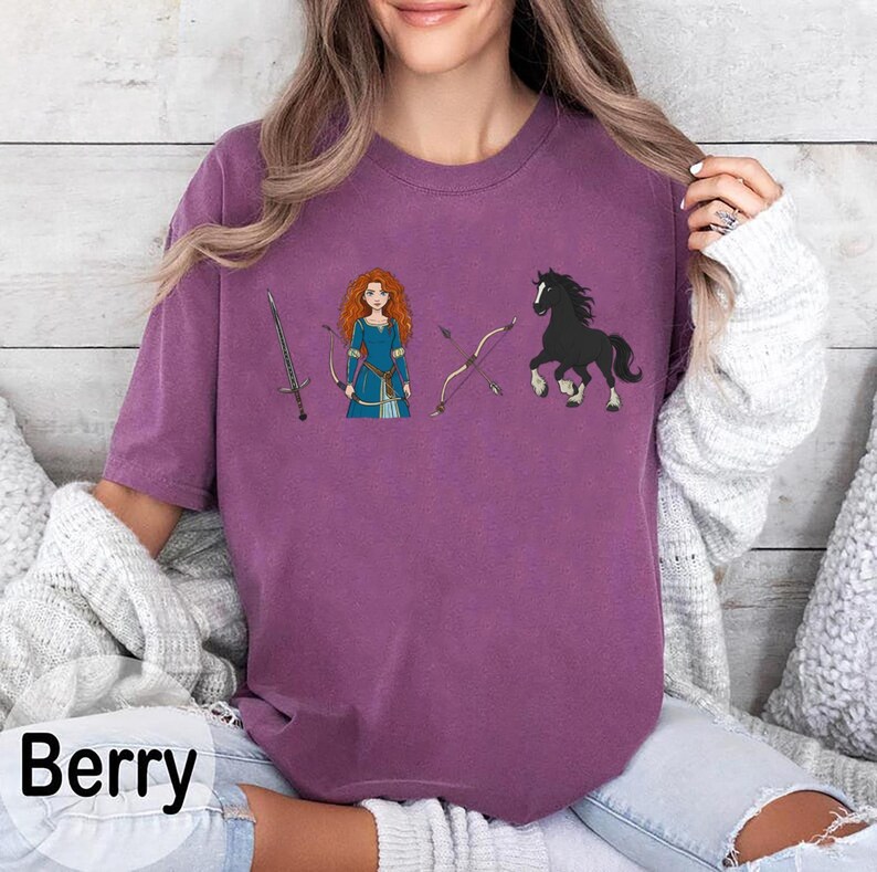 May include: Berry-colored t-shirt featuring a cartoon illustration of a character with red hair, a sword, bow and arrow, and a black horse. The shirt has a relaxed fit and short sleeves. The word "Berry" is printed in white at the bottom left.