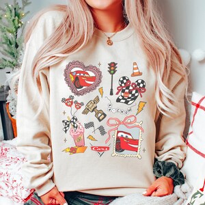May include: Beige sweatshirt featuring a colorful graphic design with cartoon cars, hearts, checkered flags, and lightning bolts. The design includes the number 95 and the word "Cars".