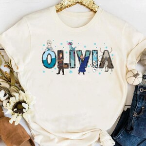 Pu&ograve; includere: T-shirt color crema con il nome "OLIVIA" in un design stravagante. Ogni lettera &egrave; decorata con personaggi e fiocchi di neve di un popolare film d'animazione. &Egrave; un capo d'abbigliamento casual.