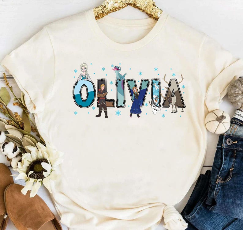 Pu&ograve; includere: T-shirt color crema con il nome "OLIVIA" in un design stravagante. Ogni lettera &egrave; decorata con personaggi e fiocchi di neve di un popolare film d'animazione. &Egrave; un capo d'abbigliamento casual.
