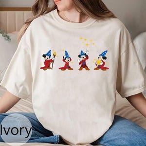 May include: Ivory-colored t-shirt featuring a graphic of Mickey Mouse in a wizard costume. The design shows Mickey in four different poses, holding a staff, pointing, and holding a star. The shirt has a crew neck and short sleeves.
