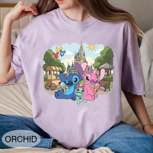 K&ouml;nnte beinhalten: Orchideenfarbenes T-Shirt mit einer Cartoon-Illustration von Stitch, Angel und einer kleinen gr&uuml;nen Kreatur, die Eiswaffeln vor einem Schloss genie&szlig;en. Das Design umfasst einen Essenswagen, B&auml;ume und Luftballons. Das Wort "ORCHID" ist unten aufgedruckt.