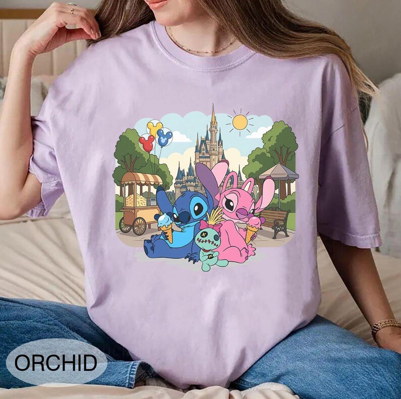 K&ouml;nnte beinhalten: Orchideenfarbenes T-Shirt mit einer Cartoon-Illustration von Stitch, Angel und einer kleinen gr&uuml;nen Kreatur, die Eiswaffeln vor einem Schloss genie&szlig;en. Das Design umfasst einen Essenswagen, B&auml;ume und Luftballons. Das Wort "ORCHID" ist unten aufgedruckt.