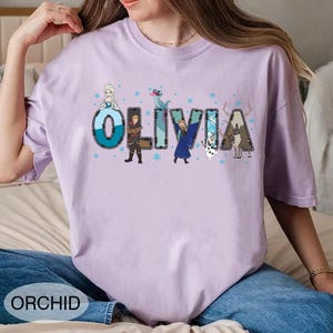 Pu&ograve; includere: Una maglietta lilla chiaro con il nome "OLIVIA" in un carattere decorativo, con personaggi dei cartoni animati e fiocchi di neve. La parola "ORCHID" &egrave; stampata in basso sulla maglietta. La maglietta &egrave; realizzata in un materiale morbido.